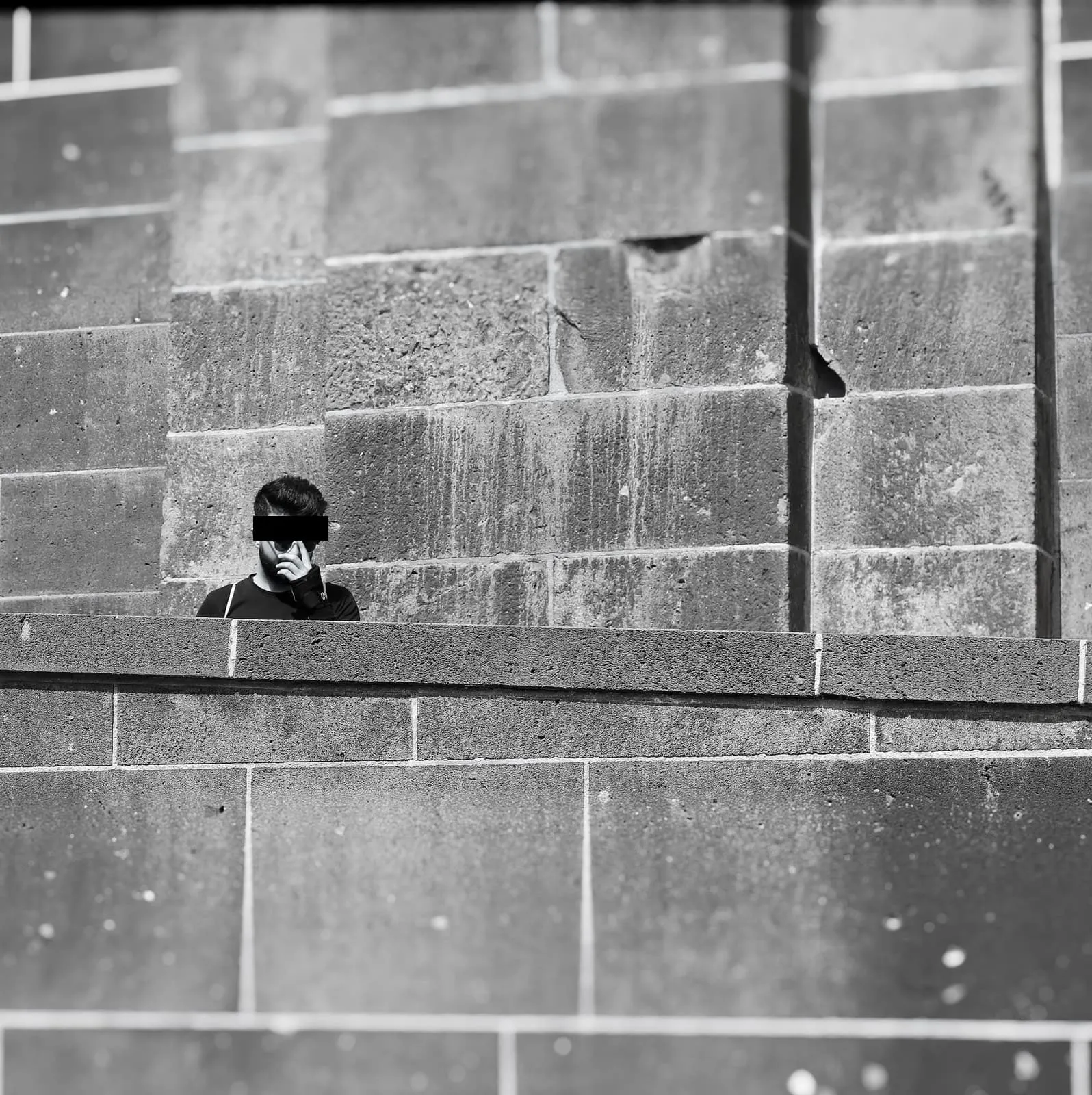 Black and white, a man behind a stone wall, hand at his face, eyes barred, 2018