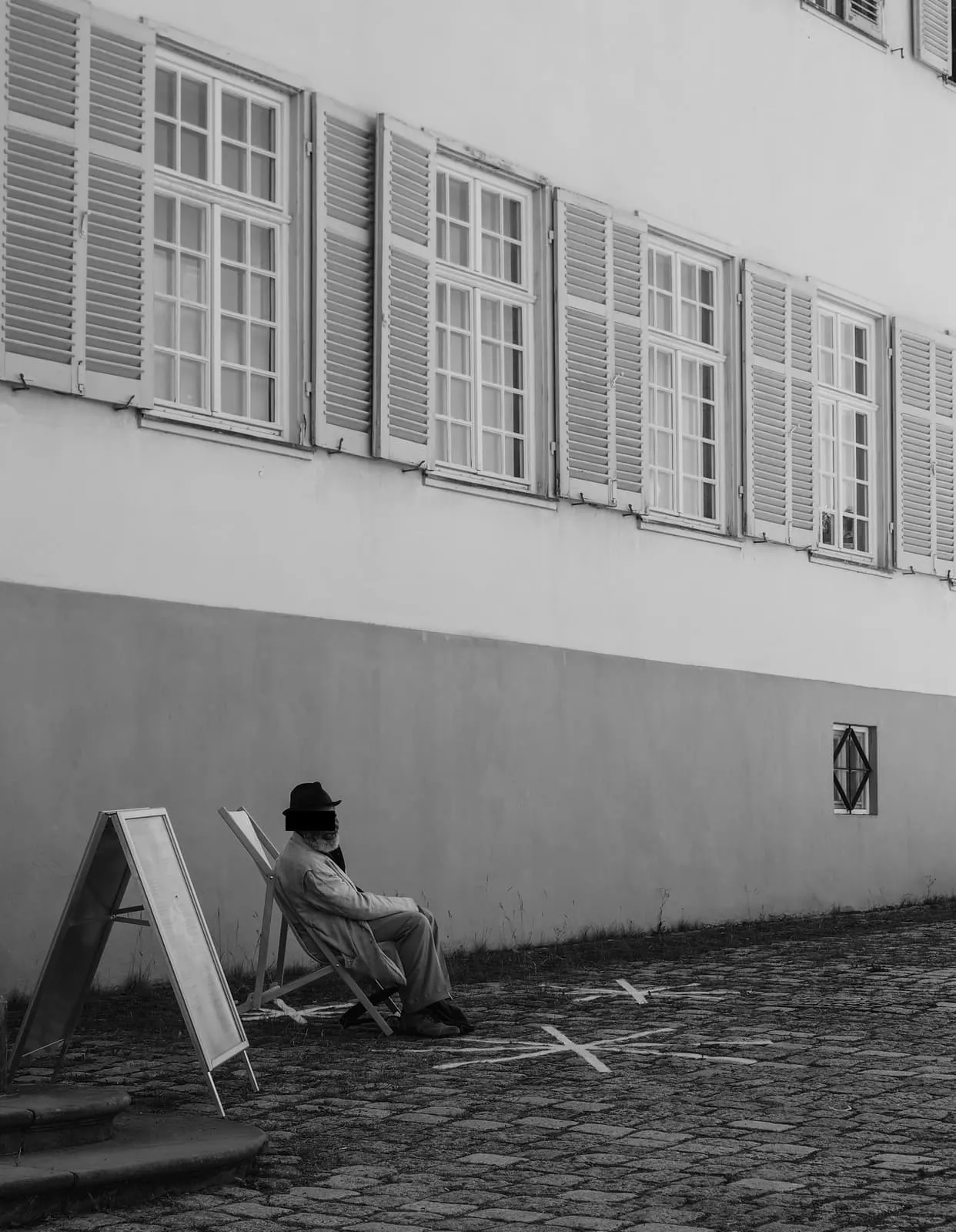 Black and white, a man in a hat on a deckchair by a long wall, eyes barred, 2019