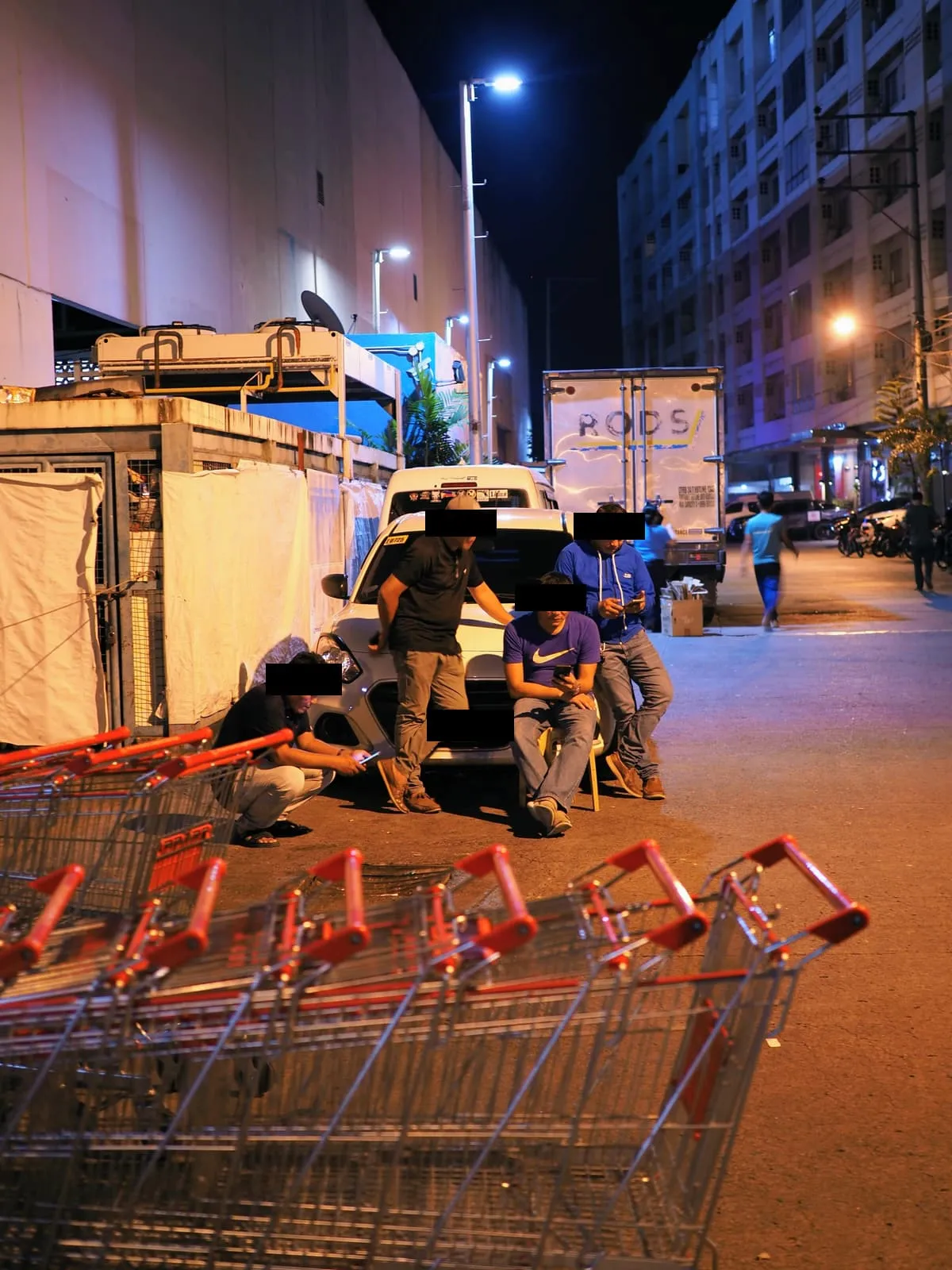Four men around a white car at night, eyes barred, trolleys in front, 2024