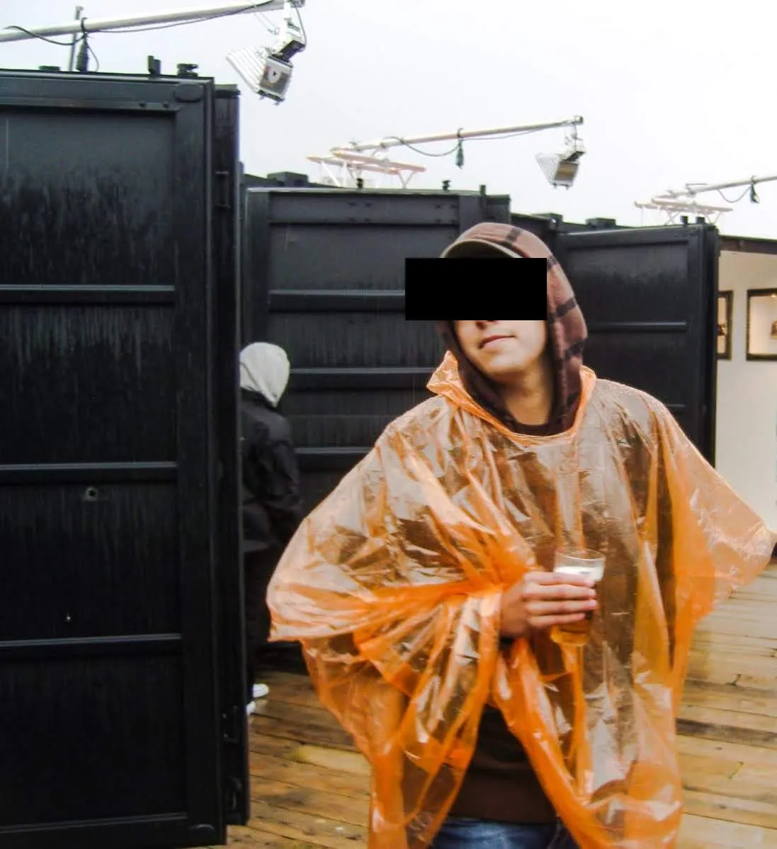 Man in an orange rain poncho holding a glass, eyes barred