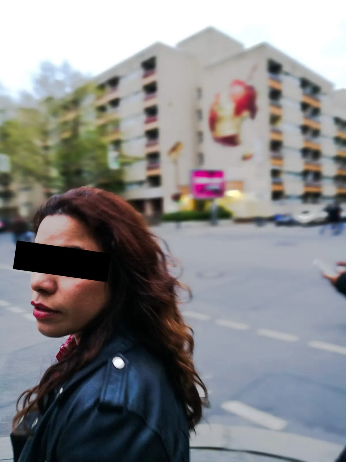 Woman in a leather jacket looking back on a street, eyes barred, 2017