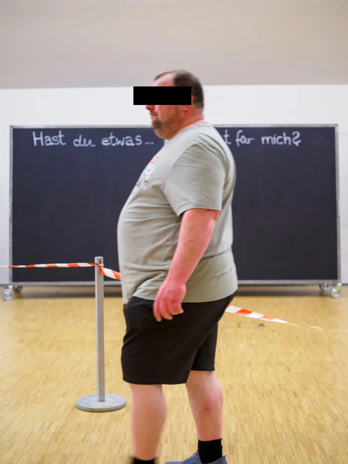 Man in profile walking past barrier tape and a blackboard, eyes barred, 2026