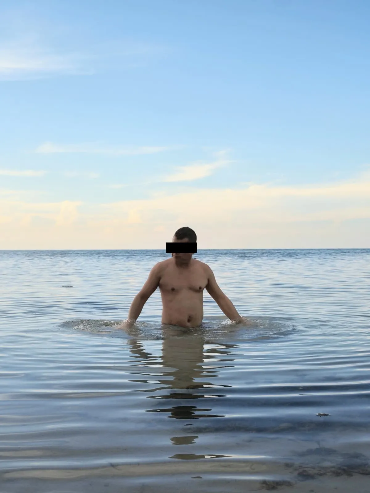 Man standing waist deep in a calm sea, eyes barred, 2024