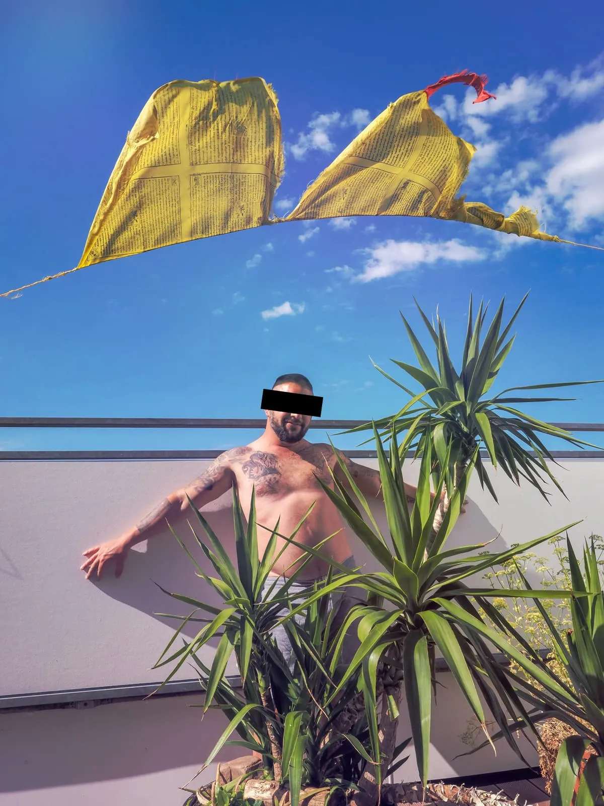 Shirtless tattooed man on a roof terrace under yellow flags, eyes barred, 2020