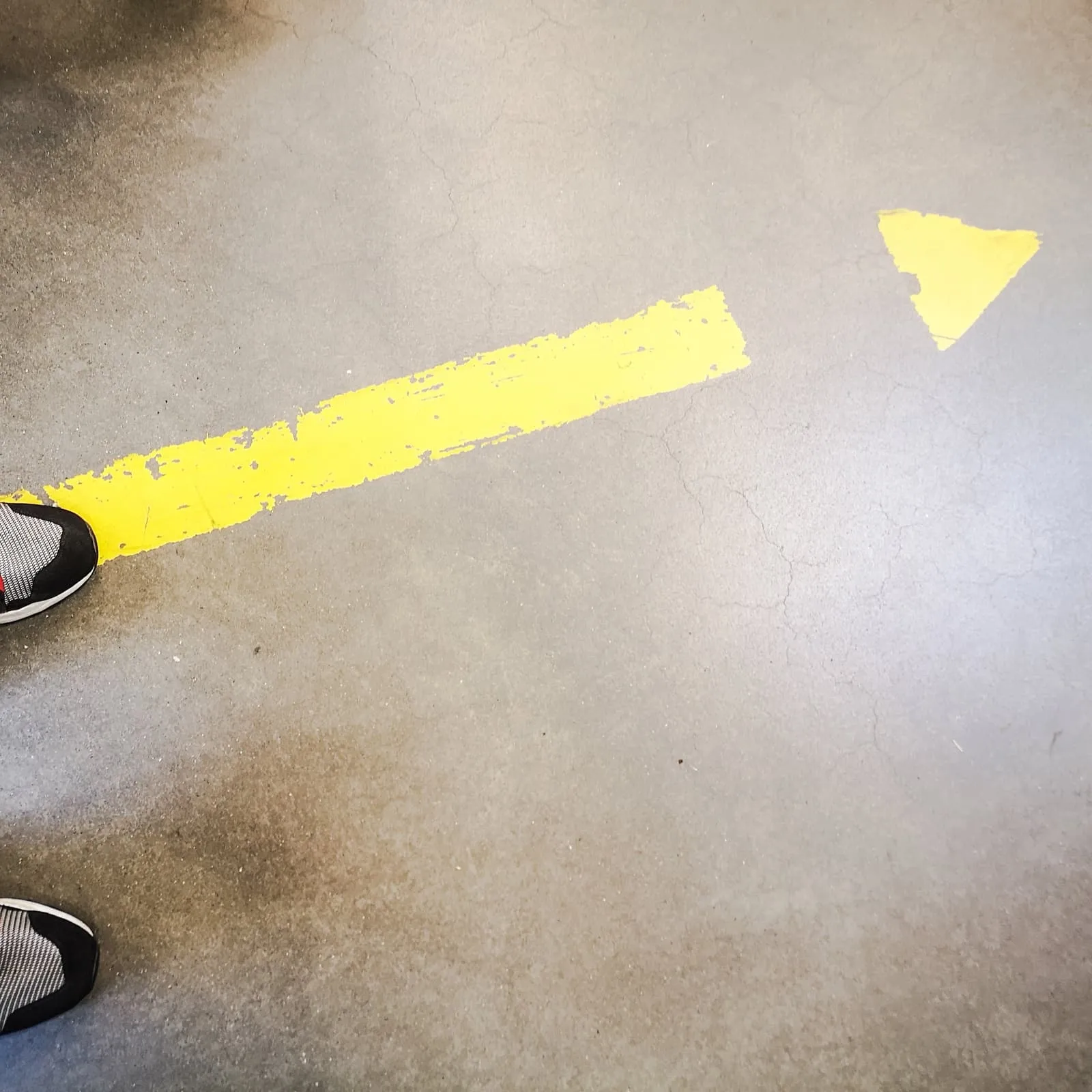 Yellow arrow painted on a floor, two shoe tips at the edge, 2019