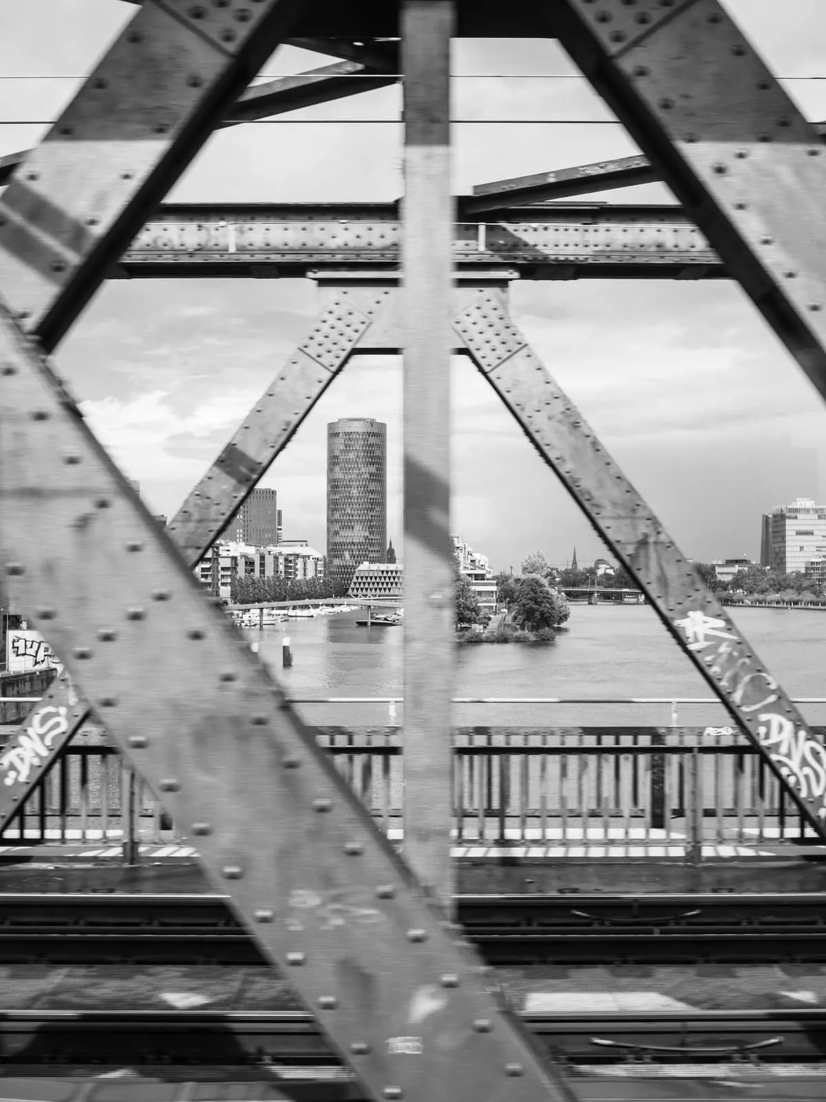 Black and white riveted bridge truss framing a river and towers, 2023