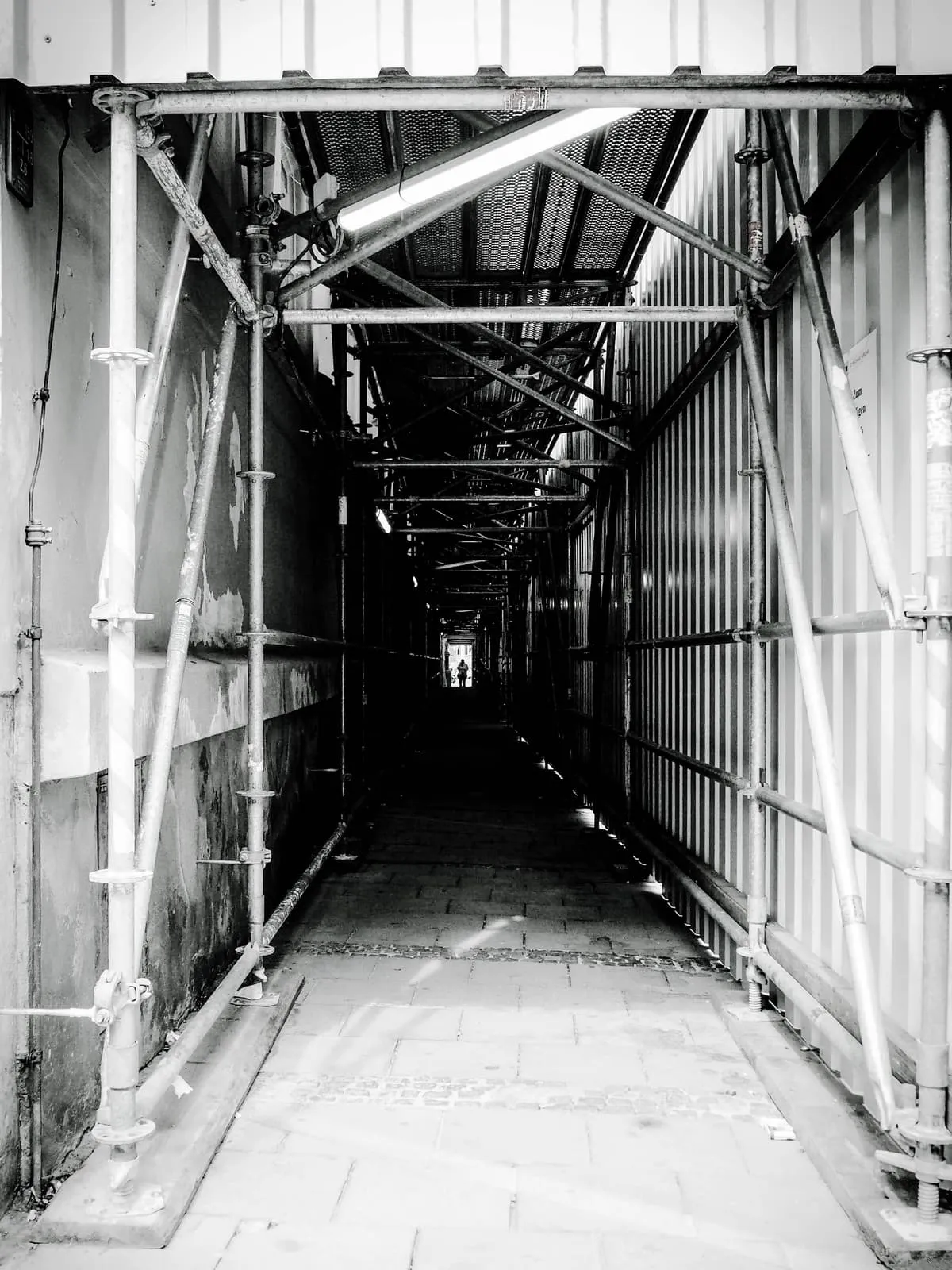 Black and white scaffolding tunnel over a pavement, a figure at the far end