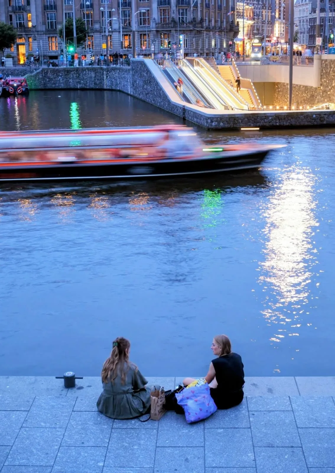 Two people sitting on a quay wall, from behind, a boat passing, 2024