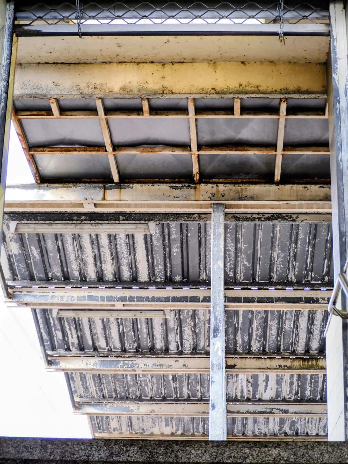 Underside of a station footbridge, corroded metal steps from below, 2025
