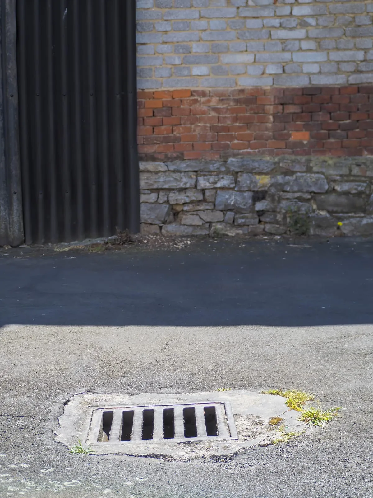 Street drain in patched asphalt before a brick wall, 2020
