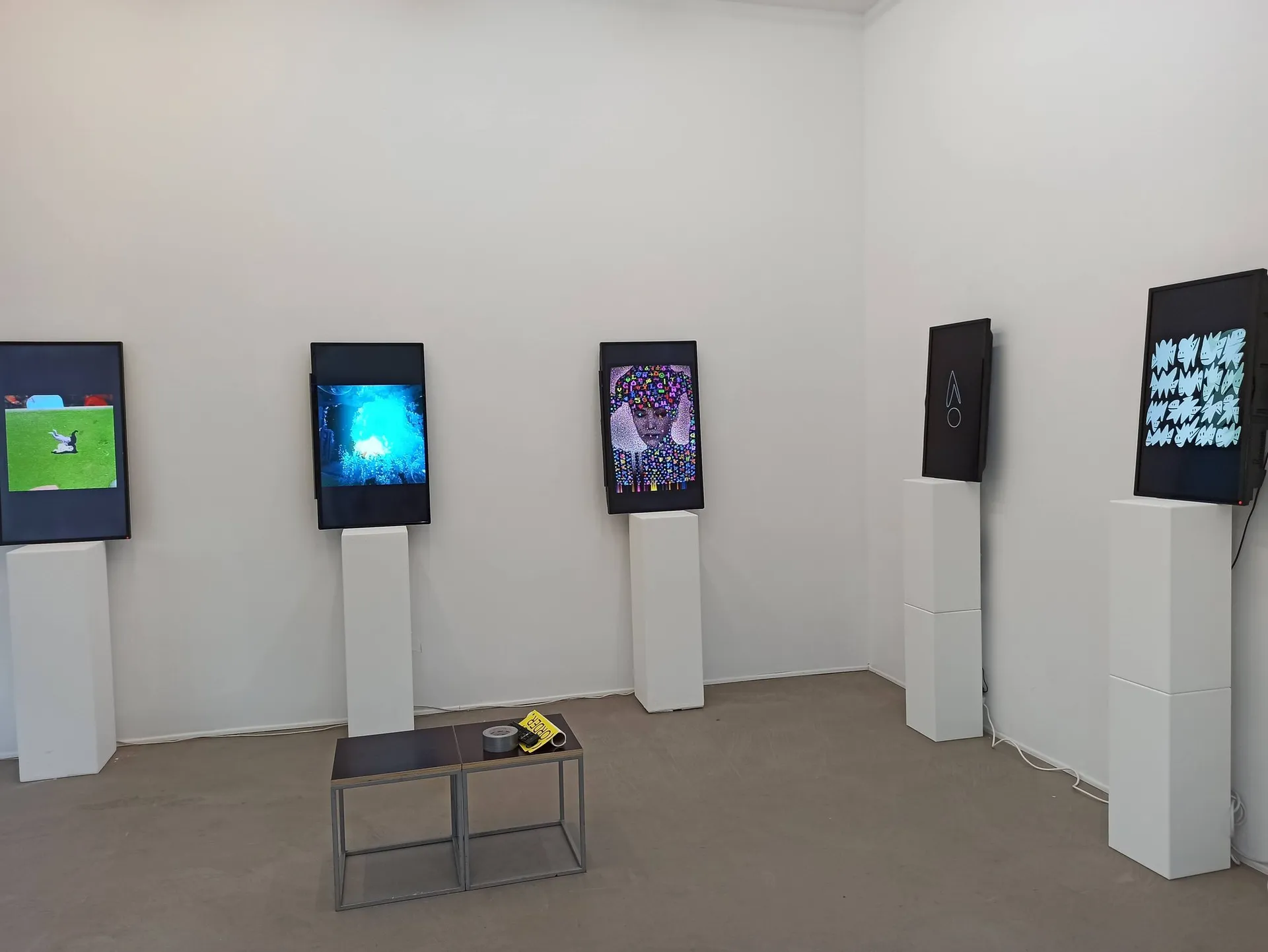 Fünf hochformatige Displays mit digitaler Kunst auf weißen Sockeln im Galerieraum